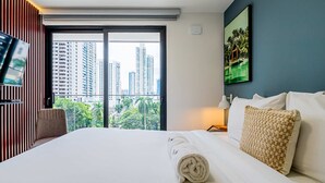 A hotel room with a large bed, a TV on the left, and a view of tall buildings through the window.