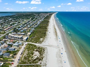 An aerial view of a coastal area with a long sandy beach, a row of residential buildings, and a clear blue ocean.
