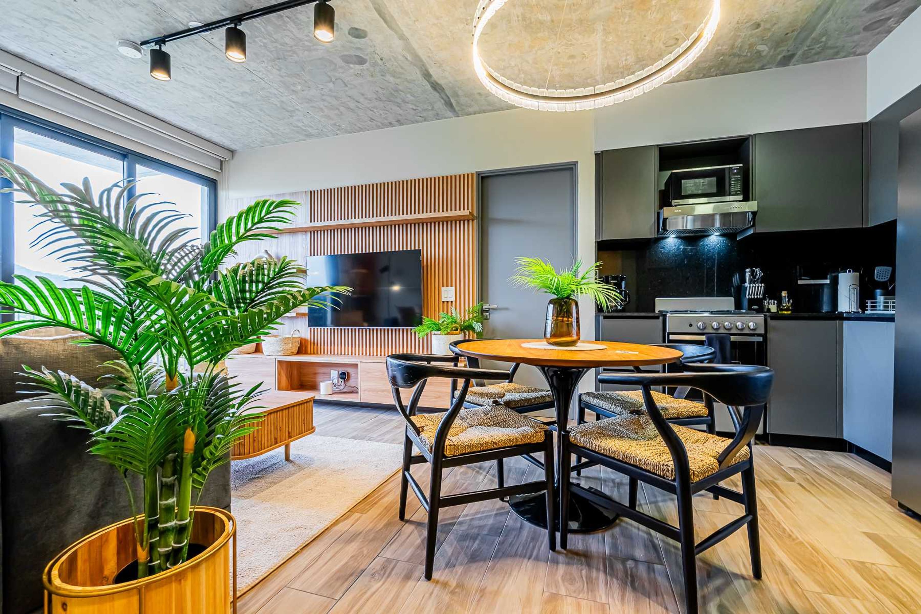 A modern living room with a round wooden table, black chairs, and a large TV. There is a kitchen area with a microwave and stove.