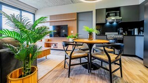 Una sala de estar moderna con una mesa redonda de madera, sillas negras y un televisor grande. Cuenta con una zona de cocina con microondas y estufa.