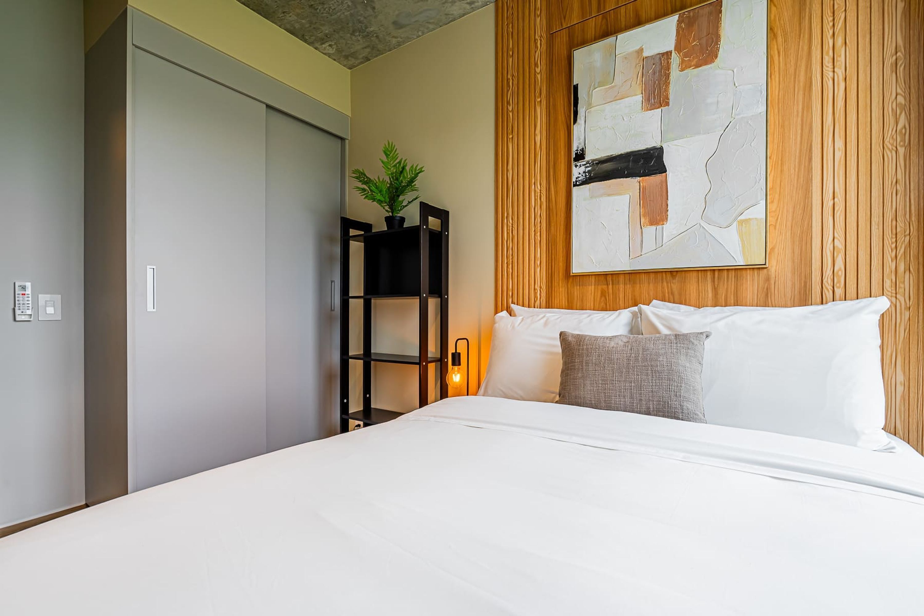 A modern bedroom with a large bed, a wooden headboard, a wall-mounted abstract painting, and a small plant on a shelf.