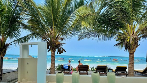 Ein Poolbereich mit Liegestühlen, einer Palme und Blick auf das Meer mit Booten.