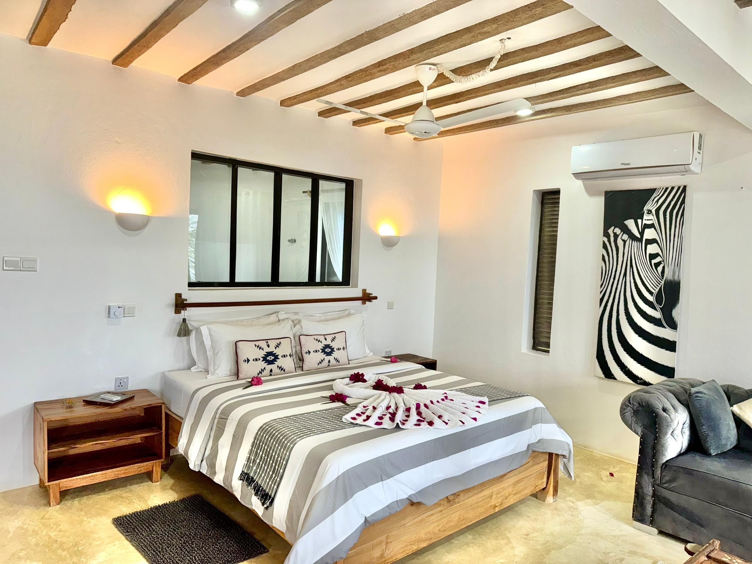 Une chambre à coucher comprenant un lit, des tables de chevet, un ventilateur de plafond, un appareil de climatisation et une œuvre d’art représentant un zèbre, accrochée au mur.