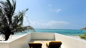 Ein Balkon mit zwei Holzstühlen und einem gelben Kissen, mit Blick auf einen Strand mit Booten und einem wolkenlosen Himmel.