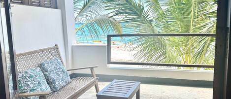 A balcony with a wicker sofa and a wooden table, overlooking a beach with palm trees and a clear blue sky.
