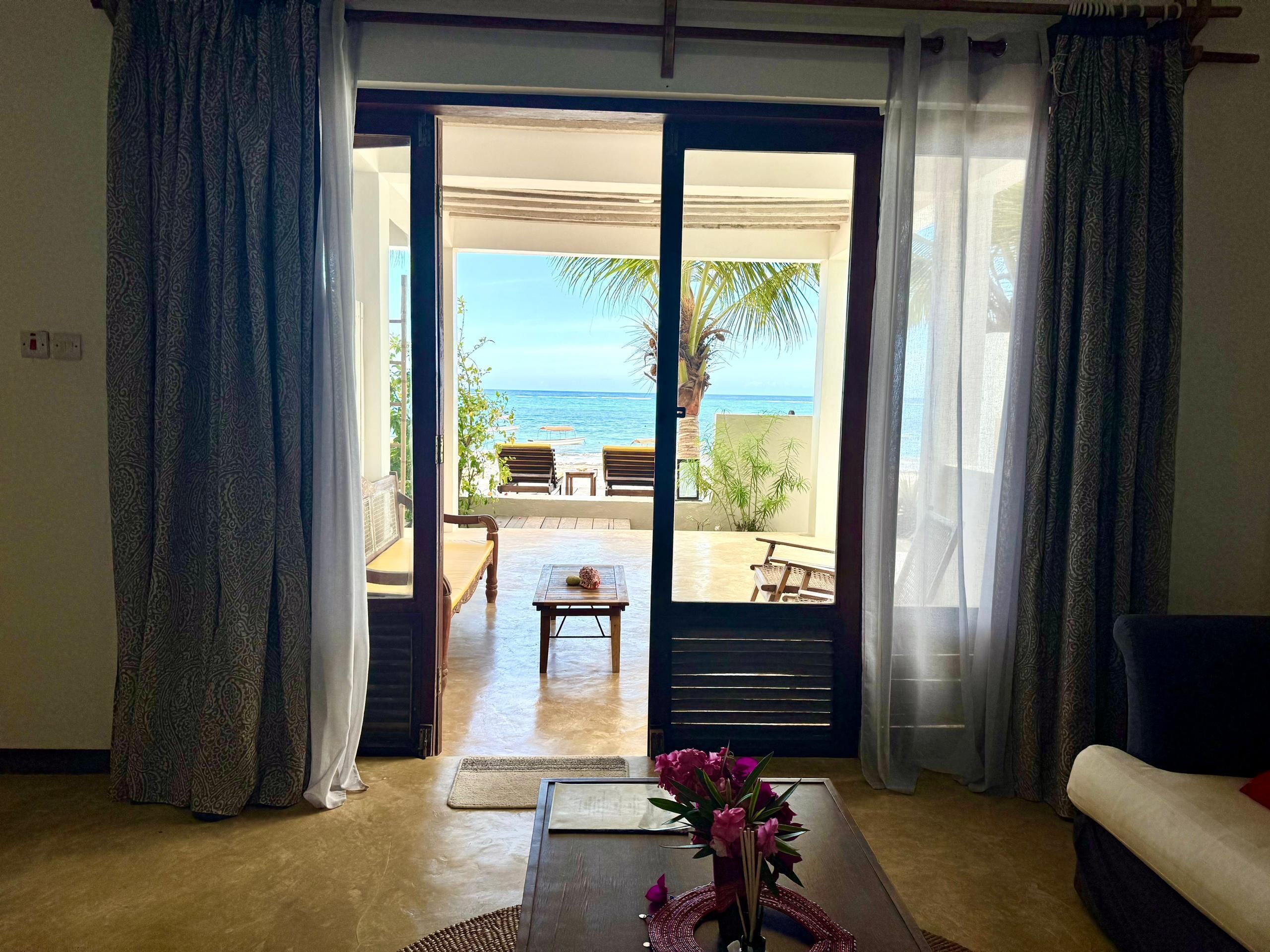 A room with a view of the sea through an open door, a wooden table with a vase of flowers, and a sofa.