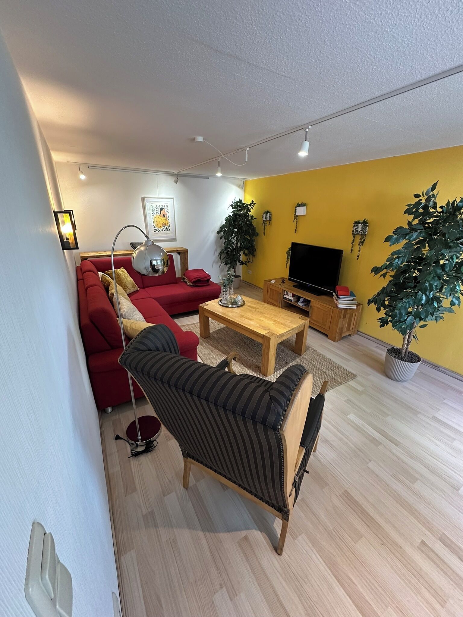 Ein modernes Wohnzimmer mit einem roten Sofa, einem gestreiften Sessel, einem Holztisch und einem Fernseher. Üchtöpfe und ein Gemälde zieren die Wand.