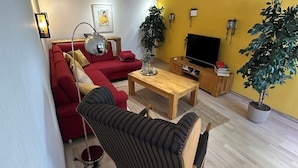 Een moderne woonkamer met een rode bank, een gestreepte fauteuil, een houten salontafel en een televisie. Er staan potplanten en er hangt een schilderij aan de muur.