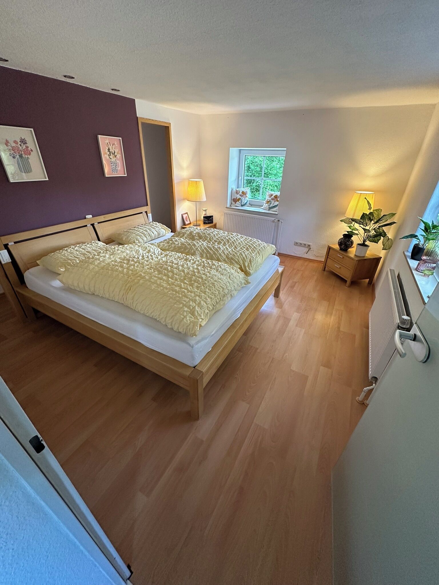 Een slaapkamer met een houten bed, een nachtkastje met een lamp, een raam met uitzicht op groen en een radiator.