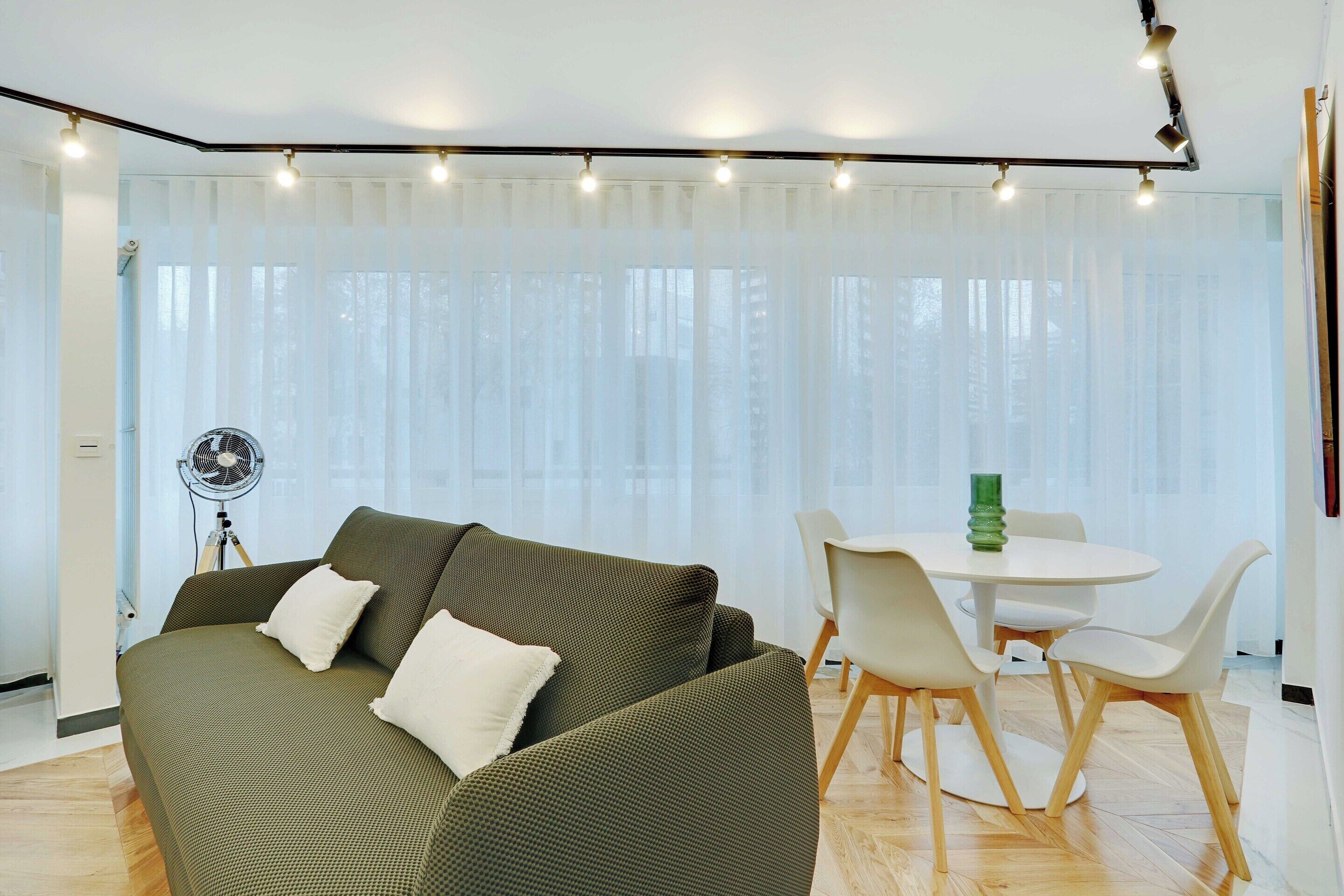 A modern living room with a sofa, a dining table with chairs, and a standing fan.