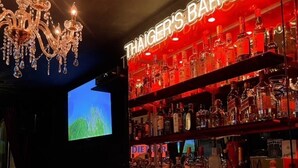 Um bar com um candelabro, prateleiras cheias de garrafas de bebidas e duas telas exibindo uma paisagem verde.
