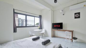 A modern hotel room with a bed, a desk with a TV displaying 'NETFLIX', a window with curtains, a small table with a chair, and a ceiling fan.