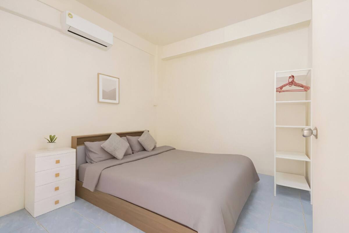 Een slaapkamer met een bed, een nachtkastje, een kleine plant, een wandairconditioner, een ingelijste foto en een kast met kledinghangers.