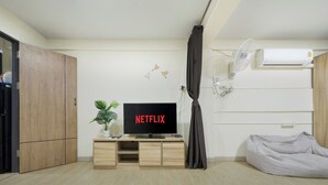 Uma sala de estar moderna com uma televisão de tela plana exibindo 'NETFLIX', um armário de madeira, uma planta em vaso, um pufe cinza e um aparelho de ar condicionado.
