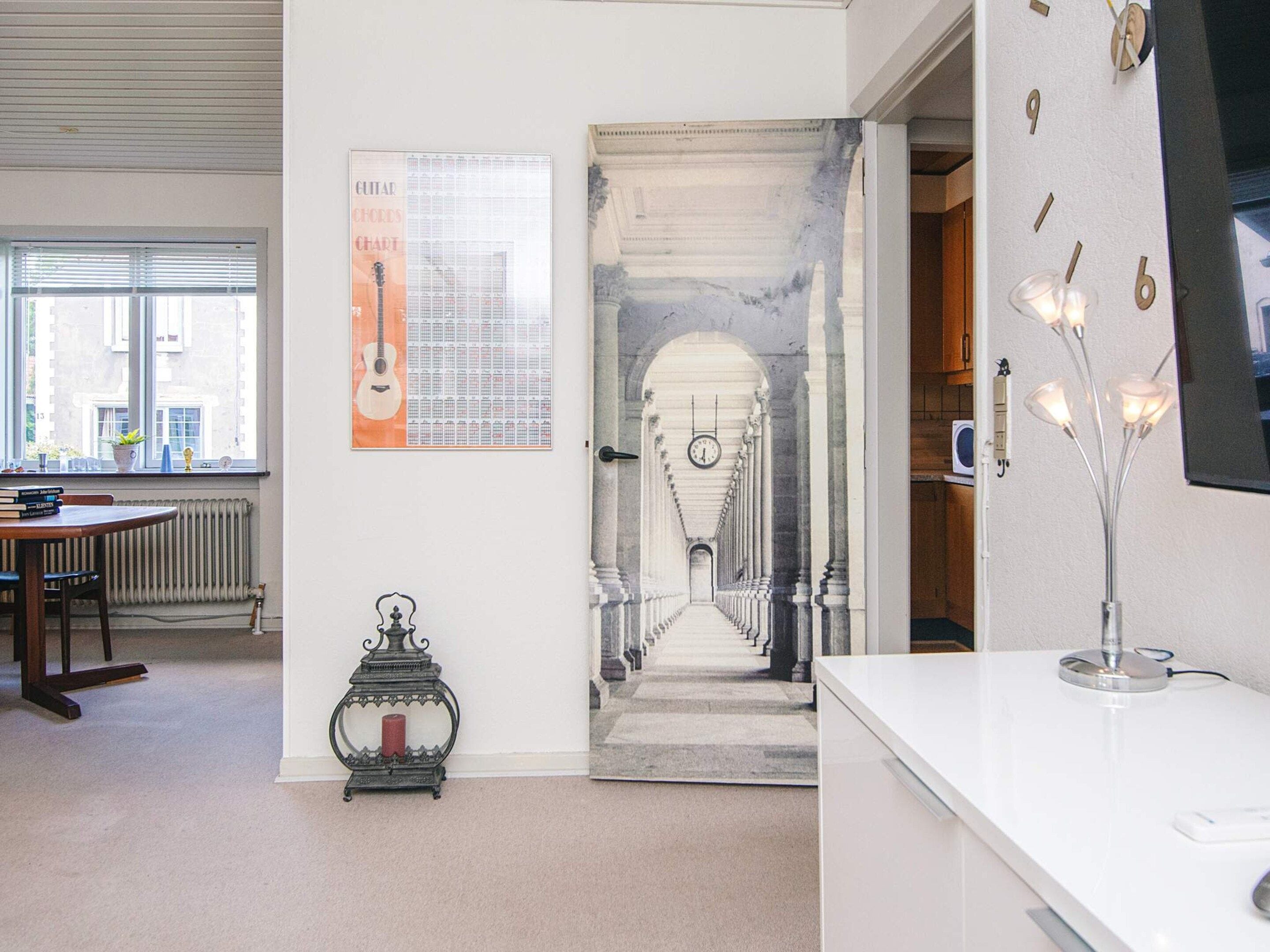 Una habitación con un escritorio blanco, un cartel con diagramas de acordes de guitarra enmarcado, un soporte metálico decorativo y vistas a un pasillo con un reloj.