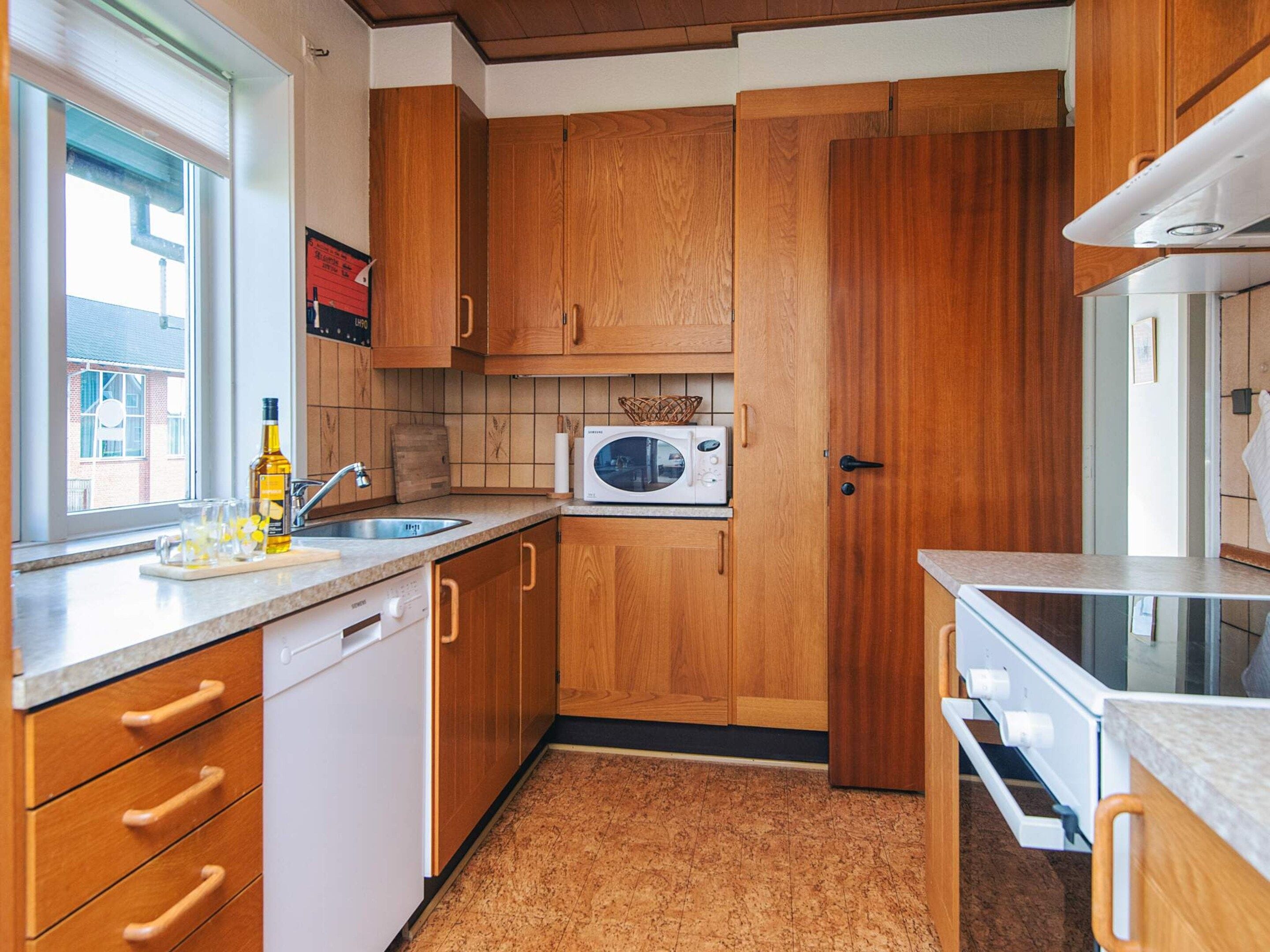 Een compacte keuken met houten kastjes, een magnetron, een vaatwasser en een raam dat voor natuurlijk licht zorgt.