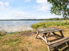 Una mesa de picnic de madera junto a un lago con orillas cubiertas de hierba y árboles al fondo.
