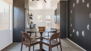 A modern kitchen with dark cabinetry, a round dining table with four chairs, and a ceiling light fixture. - Skull Rock Cottage - Walk To Town & Hot Tub, Star Gazing 1 Bedroom Home (Joshua Tree)