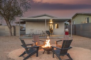 A backyard with a fire pit surrounded by Adirondack chairs, a covered patio area, and a house with a visible door and windows.