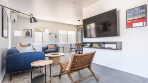 A modern living room with a blue sofa, a wooden coffee table, a wicker chair, and a flat-screen TV mounted on the wall. - Skull Rock Cottage - Walk To Town & Hot Tub, Star Gazing 1 Bedroom Home (Joshua Tree)