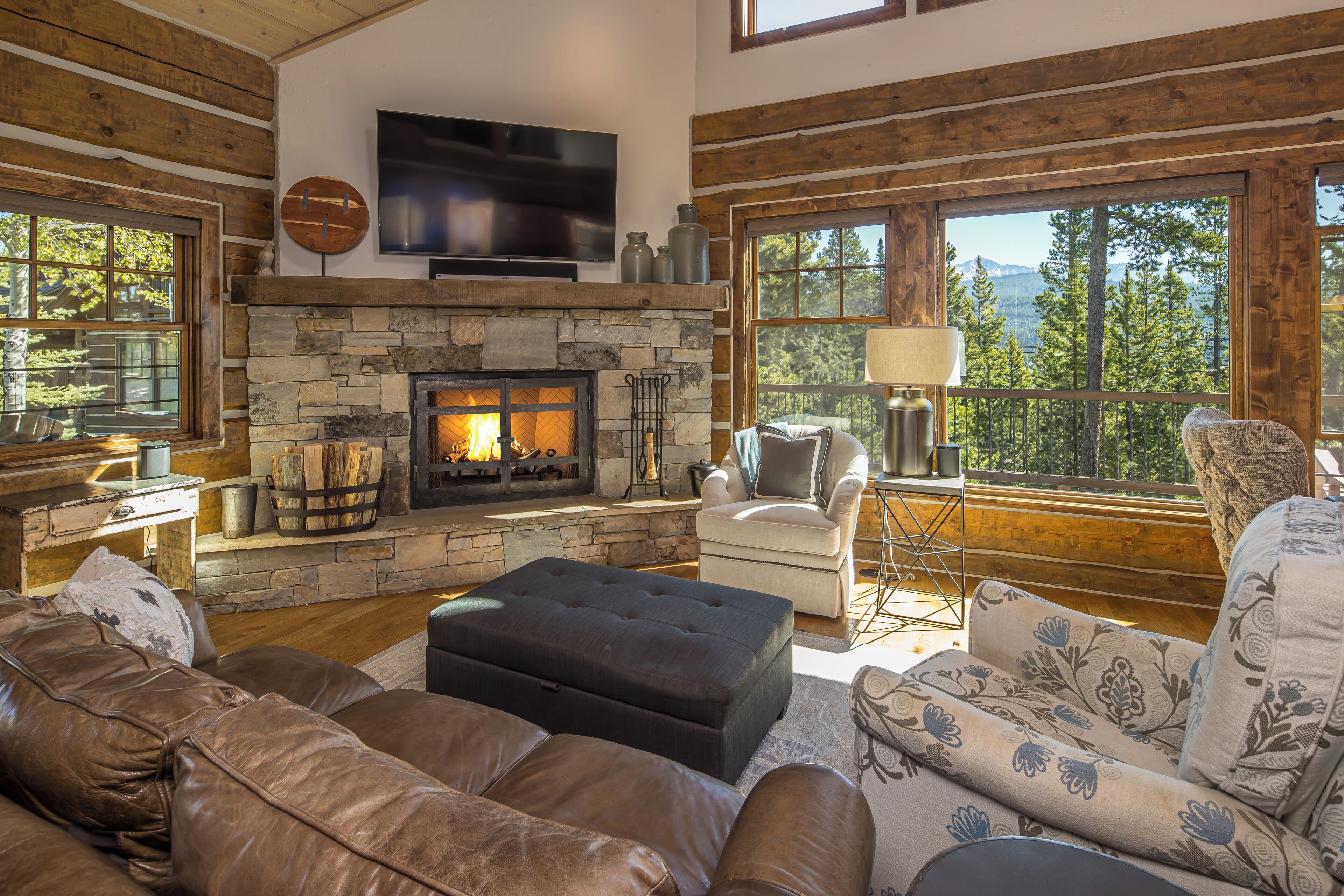 A cozy living room with a stone fireplace, leather sofas, a flat-screen TV, and large windows offering a view of trees and mountains.