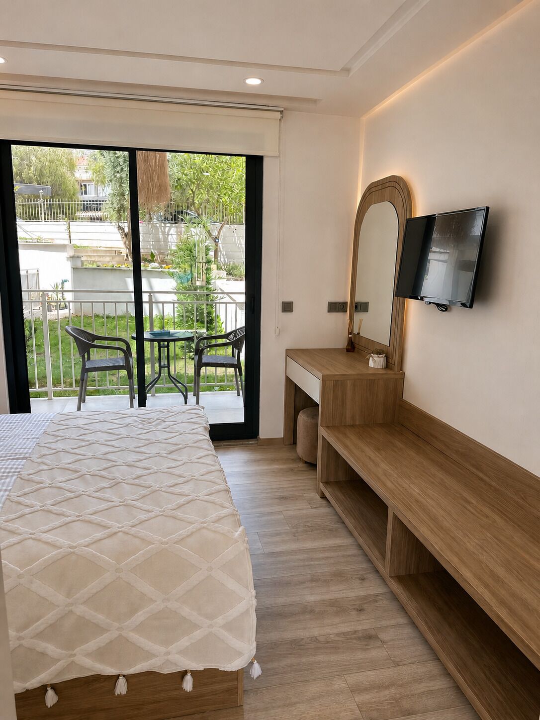 A hotel room with a bed, a TV, a bench, a mirror, and a balcony with chairs.