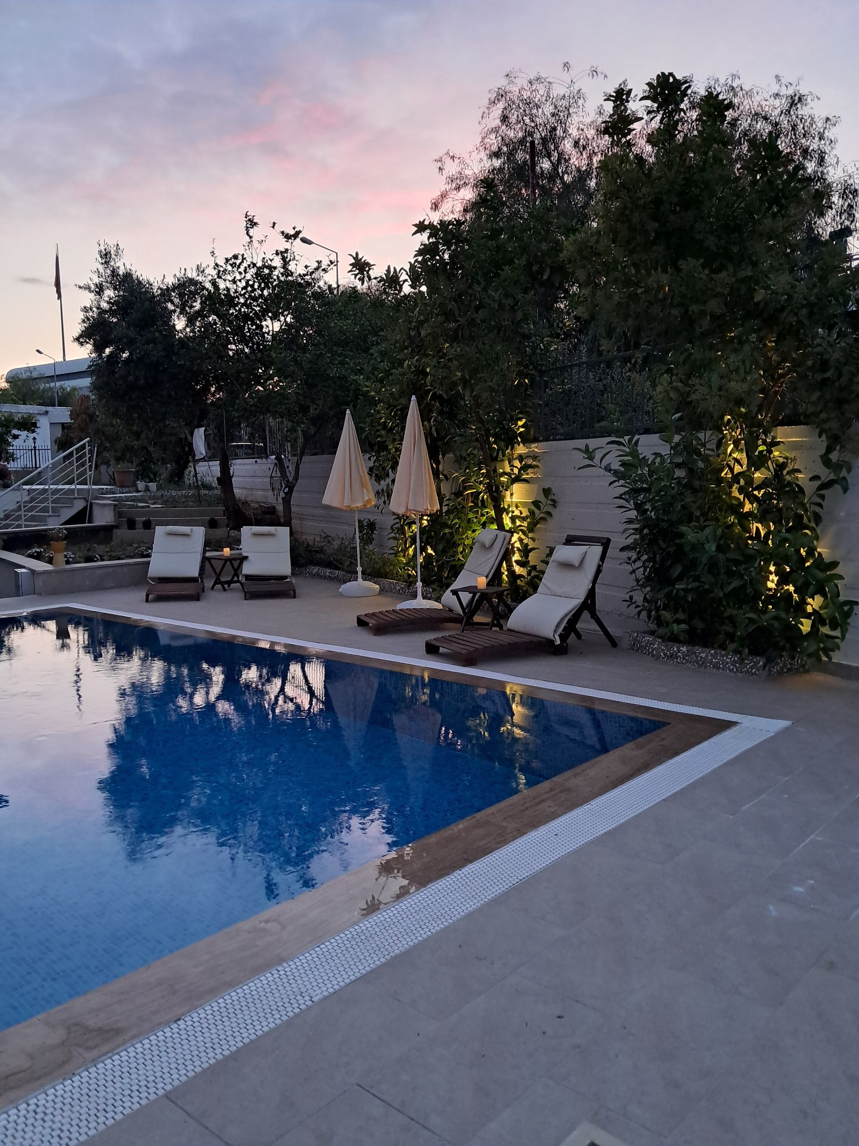 Piscina con sillones de descanso y sombrillas, rodeada de vegetación y una escalera.