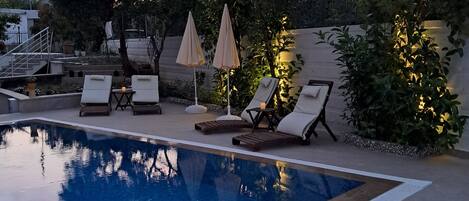 Piscina con sillones de descanso y sombrillas, rodeada de vegetación y una escalera.