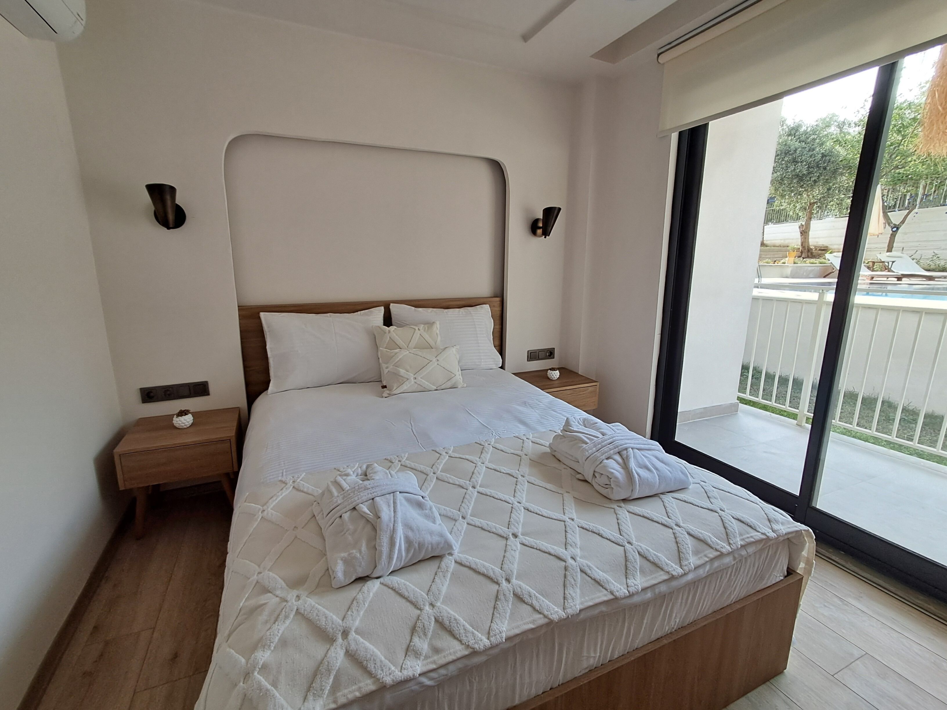 Habitación de hotel con una cama grande, cabecera de madera, ropa de cama blanca, dos mesitas de noche y vistas al exterior a través de una puerta corrediza de vidrio.