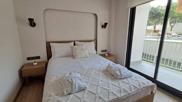 A hotel room with a large bed, wooden headboard, white bedding, two bedside tables, and a view of the outdoors through a sliding glass door.
