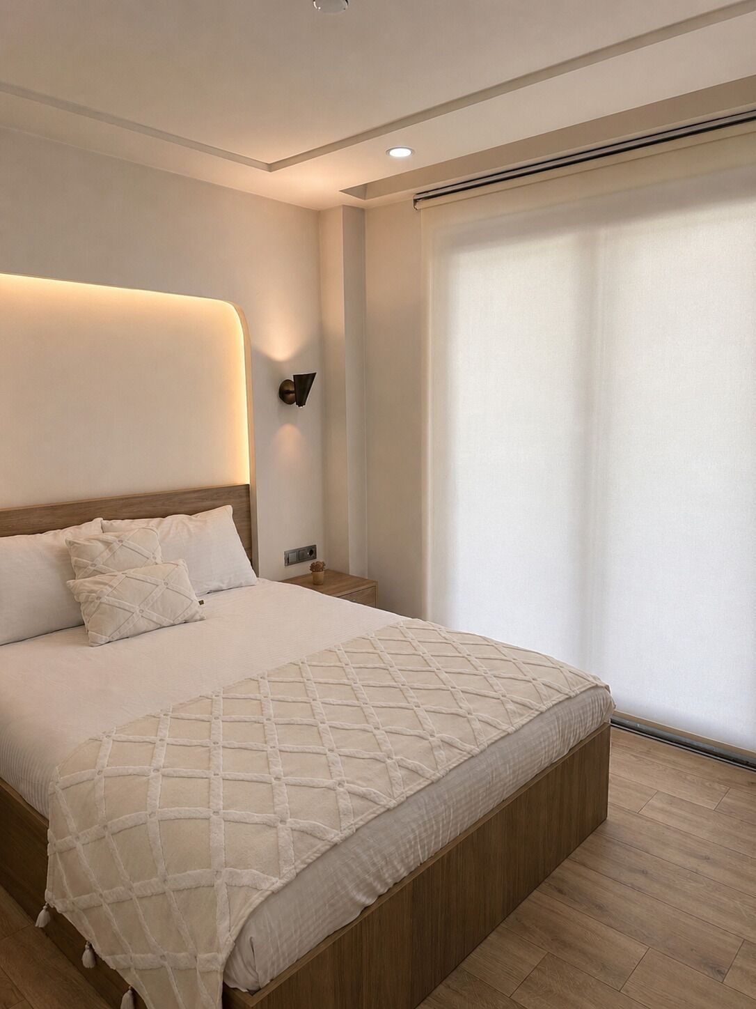 A modern bedroom with a large bed, a bedside table, a wall-mounted light, and a window with blinds.