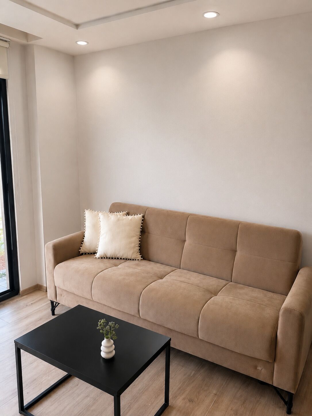 A beige sofa with two pillows, a black coffee table with a plant, and a window with blinds.