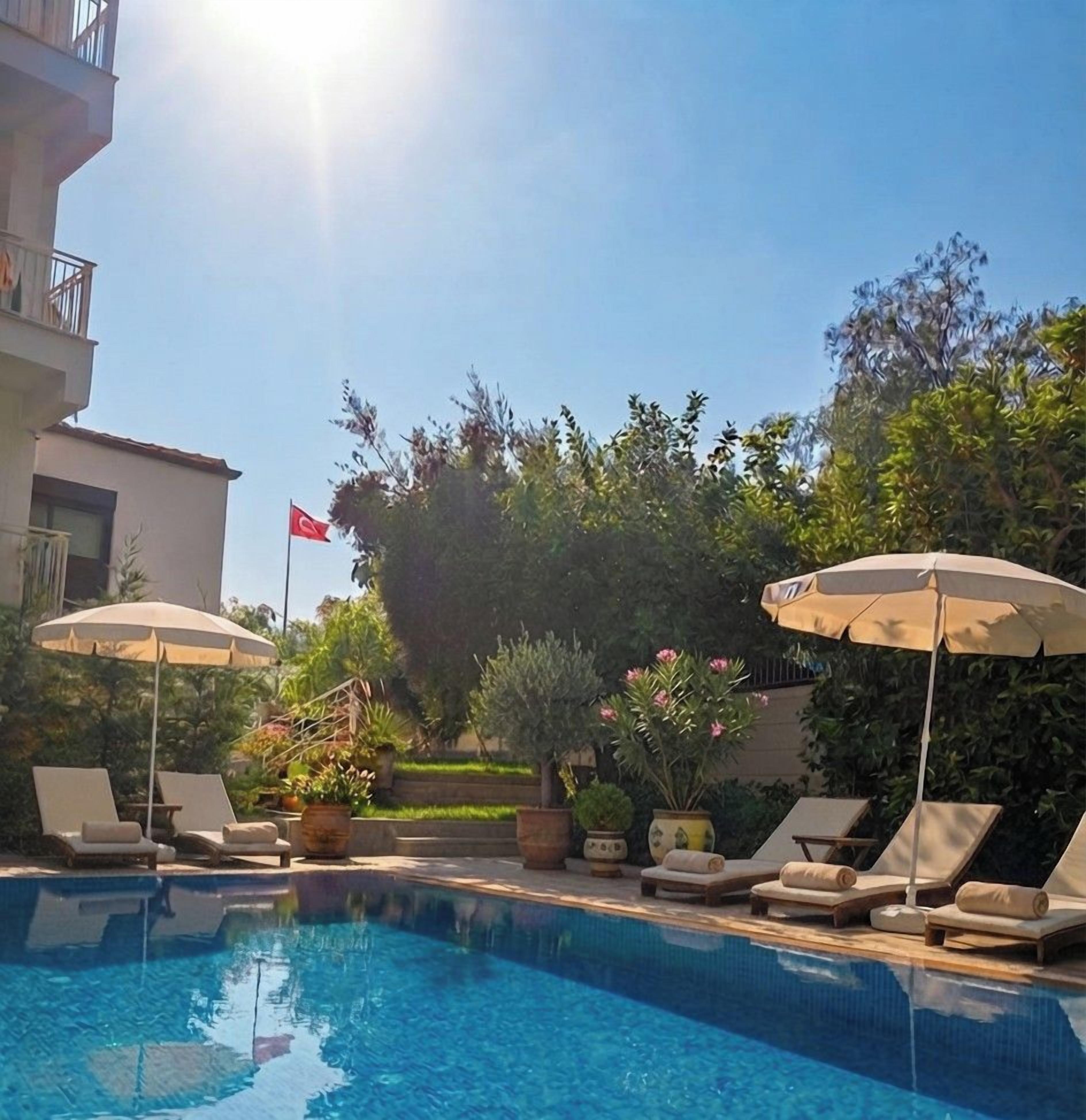 Piscina con sillas de descanso y sombrillas, rodeada de vegetación y una bandera.