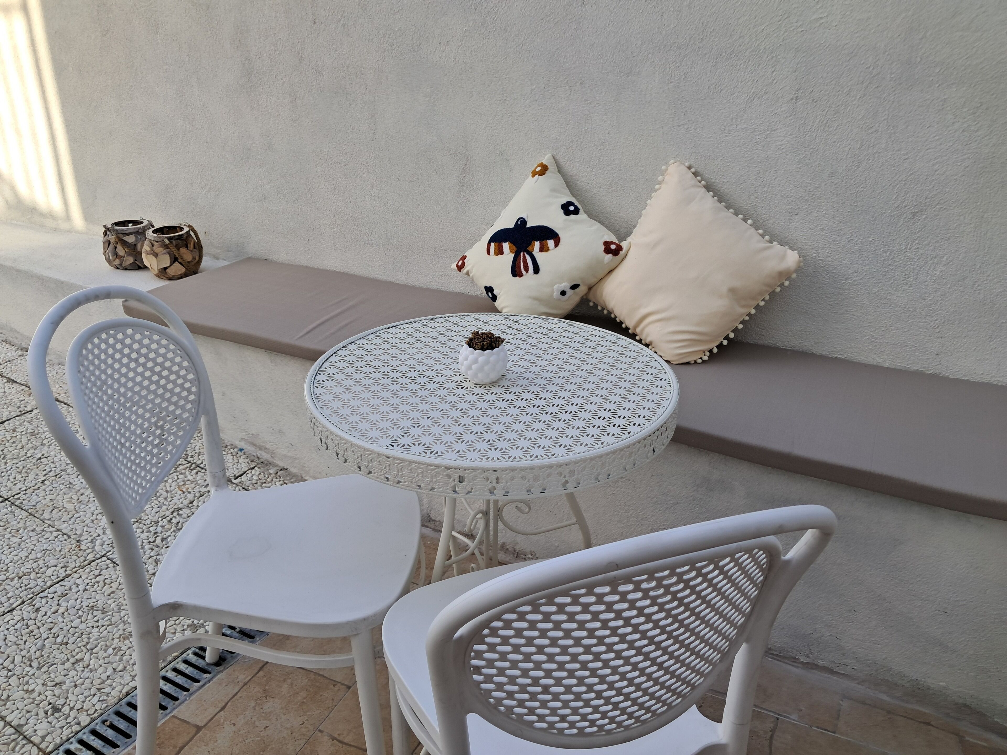 A small outdoor seating area with a round table and two white chairs, a bench with cushions, and decorative pillows.