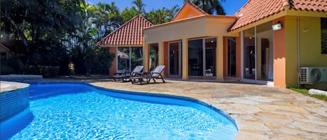 A swimming pool with a tiled edge, a covered patio area with lounge chairs, and a building with a tiled roof.