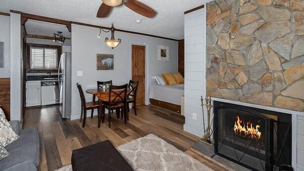 A cozy living room with a fireplace, wooden flooring, a dining area, and a kitchen visible through an open door.