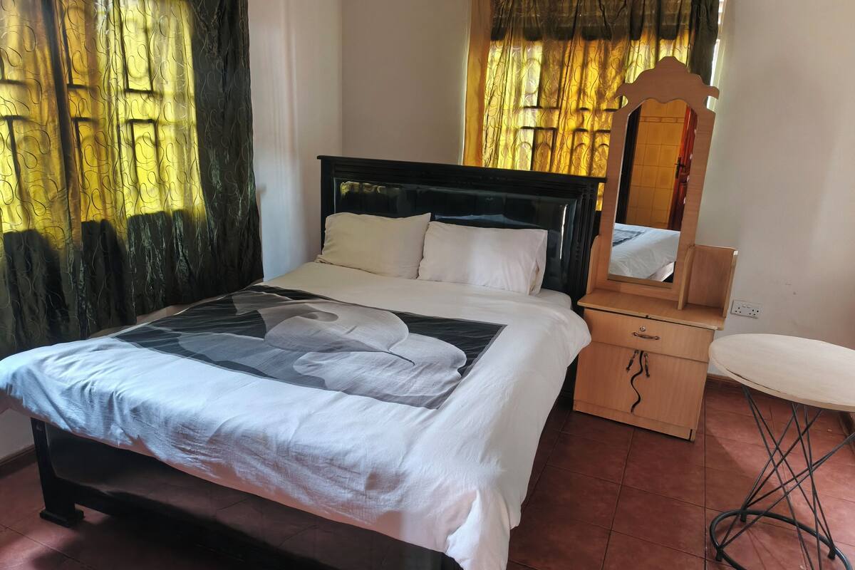 A neatly arranged bedroom with a bed, bedside table, mirror, and a small stool.