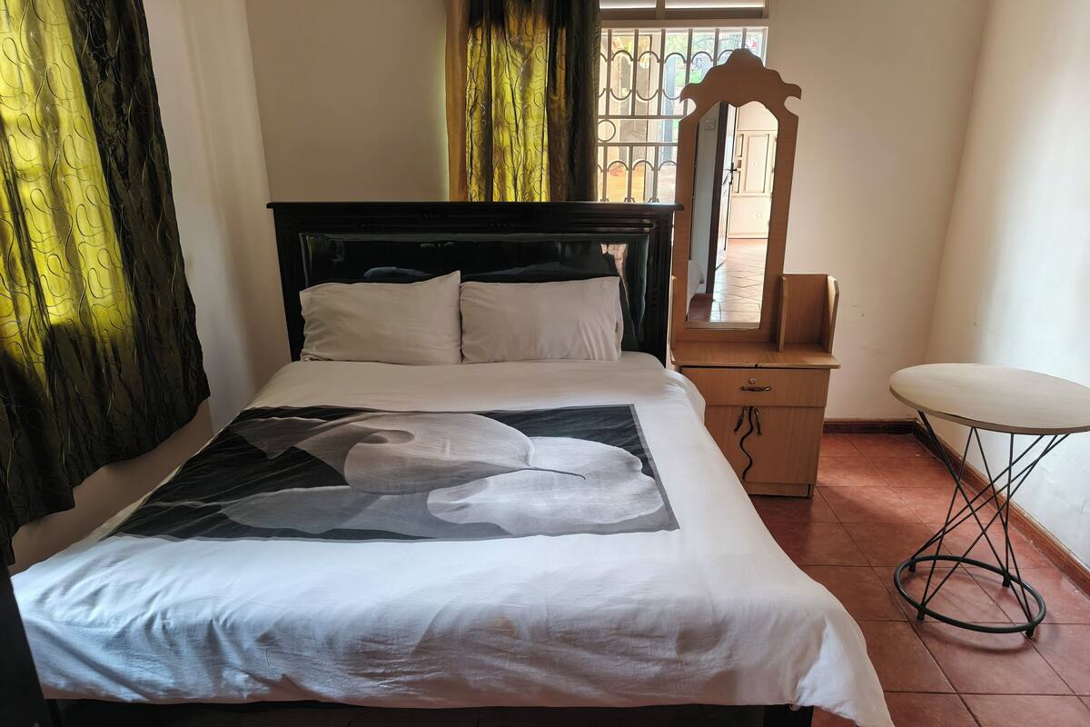 A bed with a white bedspread and a large abstract print, a wooden nightstand, a round table, and a mirror.