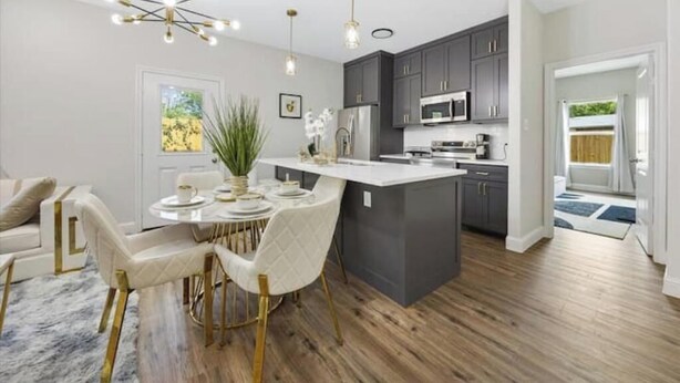 A modern kitchen with dark cabinets, a central island, and a dining area with a white table and chairs. - Homely Haven (Fort Worth)
