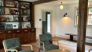A cozy living room with a wooden bookshelf, green upholstered chairs, a round wooden table, and a rustic wooden beam. - Copper Kettle Farm - newly renovated farmhouse with spectacular views (Sheffield)