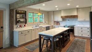 A modern kitchen with white cabinets, a central island with stools, a wooden ceiling, and a window with a stained glass light fixture. - Copper Kettle Farm - newly renovated farmhouse with spectacular views (Sheffield)
