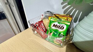 A clear glass container holding various food items, including a green packet of Nestle Milo, a jar of sugar, and several red packets, possibly snacks, on a wooden surface. - Gorgeous Modern Condo in Bacolod City (Bacolod)