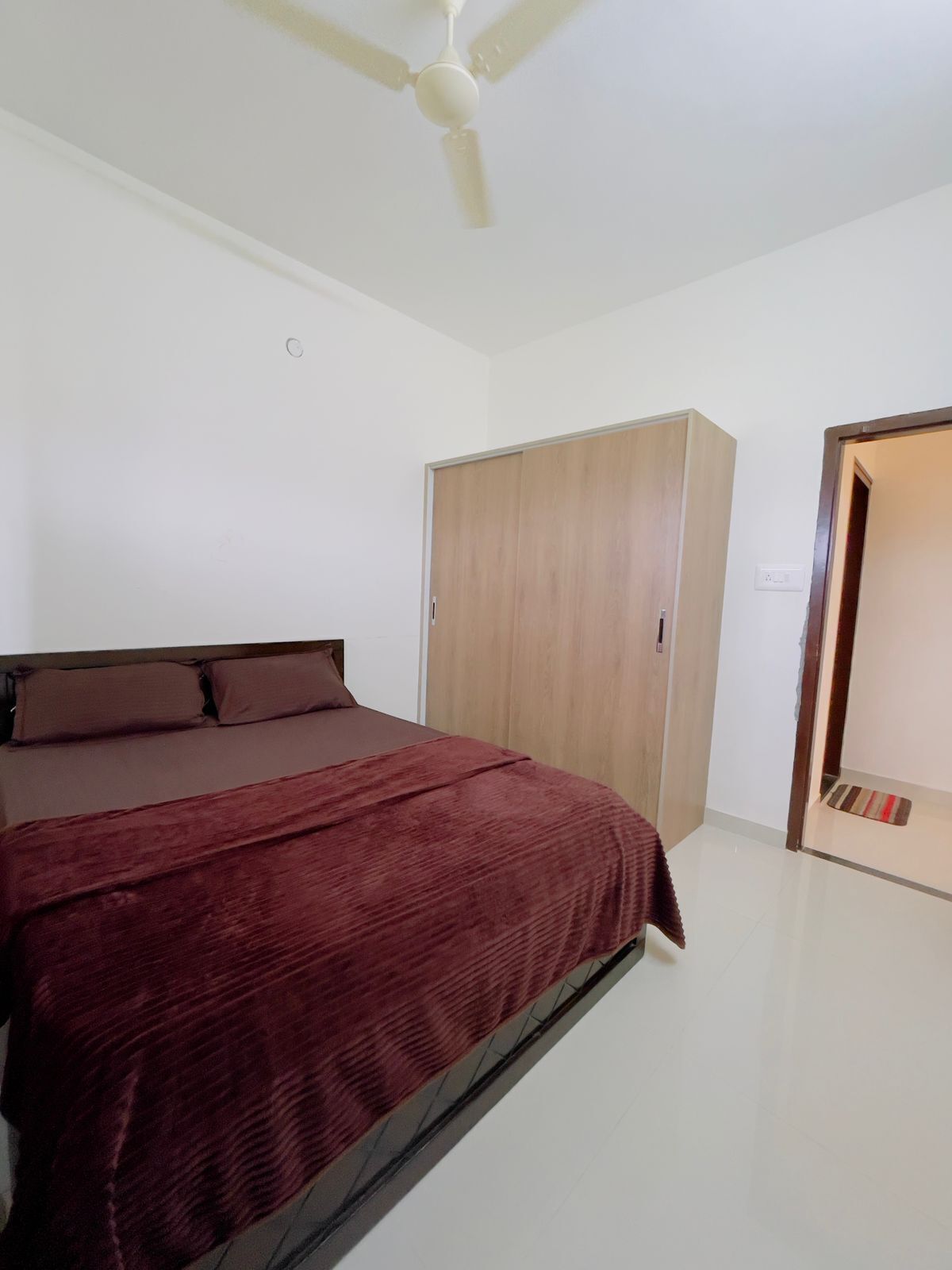 A bedroom with a bed, a wardrobe, a ceiling fan, and a door leading to another room.