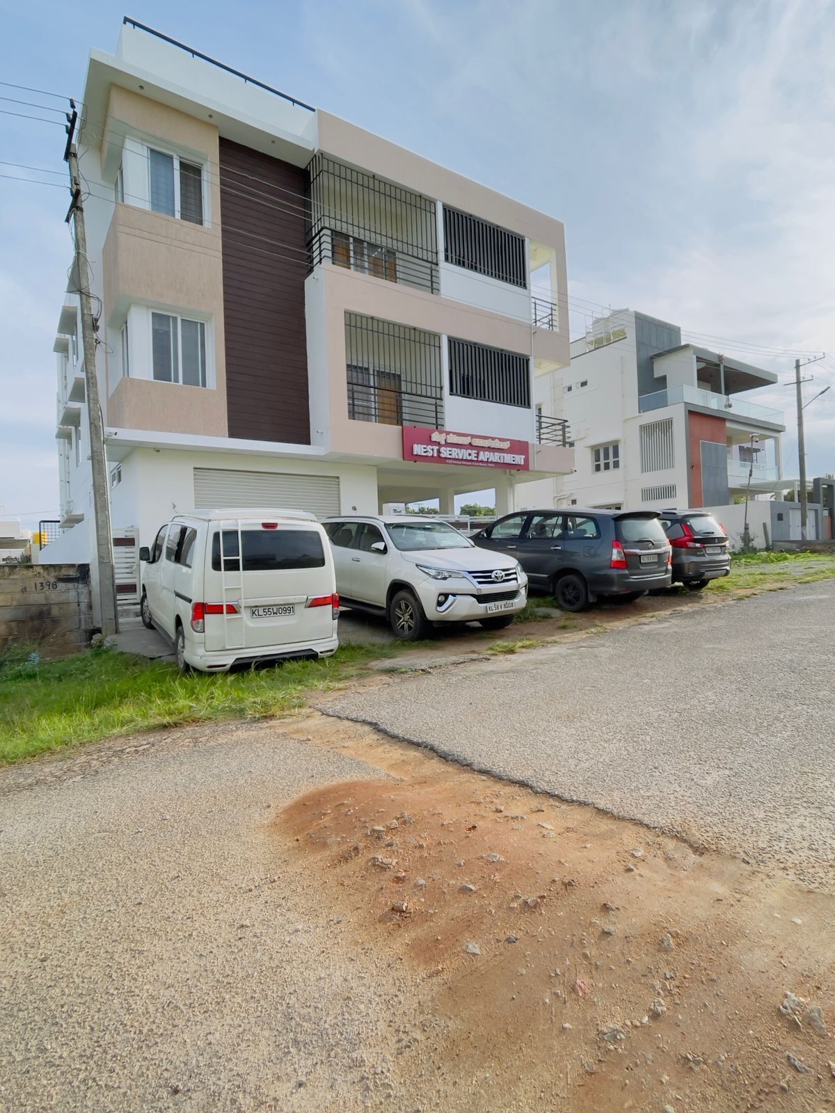 Un edificio moderno de apartamentos con un letrero que indica que es un 'West Service Apartment'. Hay varios coches estacionados en la parte delantera, y el edificio tiene un techo plano con un balcón en el piso superior.