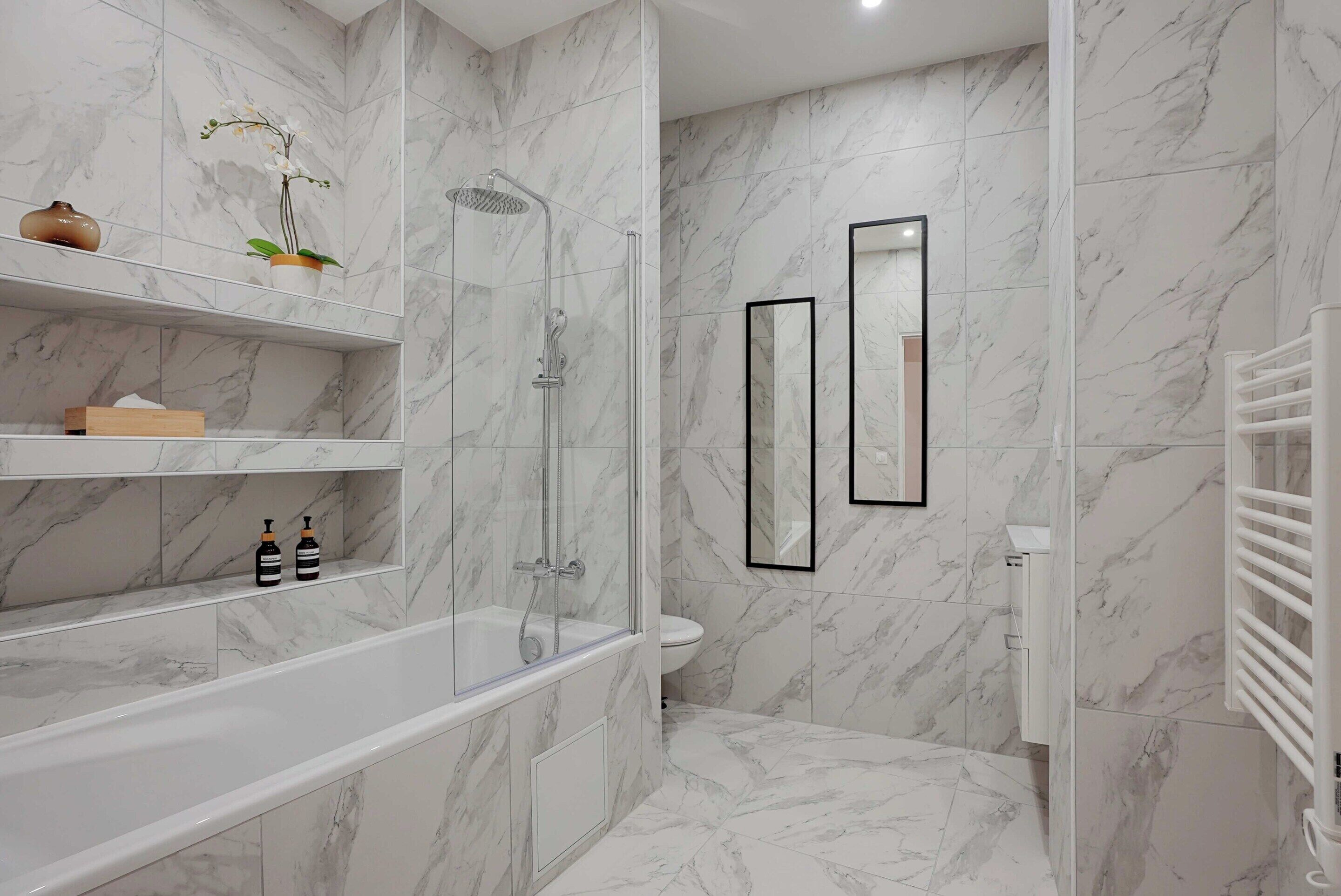 A modern bathroom with a marble floor, a glass-enclosed shower, a white bathtub, and a toilet. There are shelves with toiletries and a mirror on the wall.