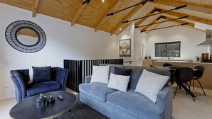 Una sala moderna con un sofá y un sillón azules, un espejo redondo y una zona de cocina con ventanal.