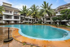 A swimming pool surrounded by a multi-story building with balconies, palm trees, and a tiled deck area. - Goan Hideaway - Cuatro (Arpora)