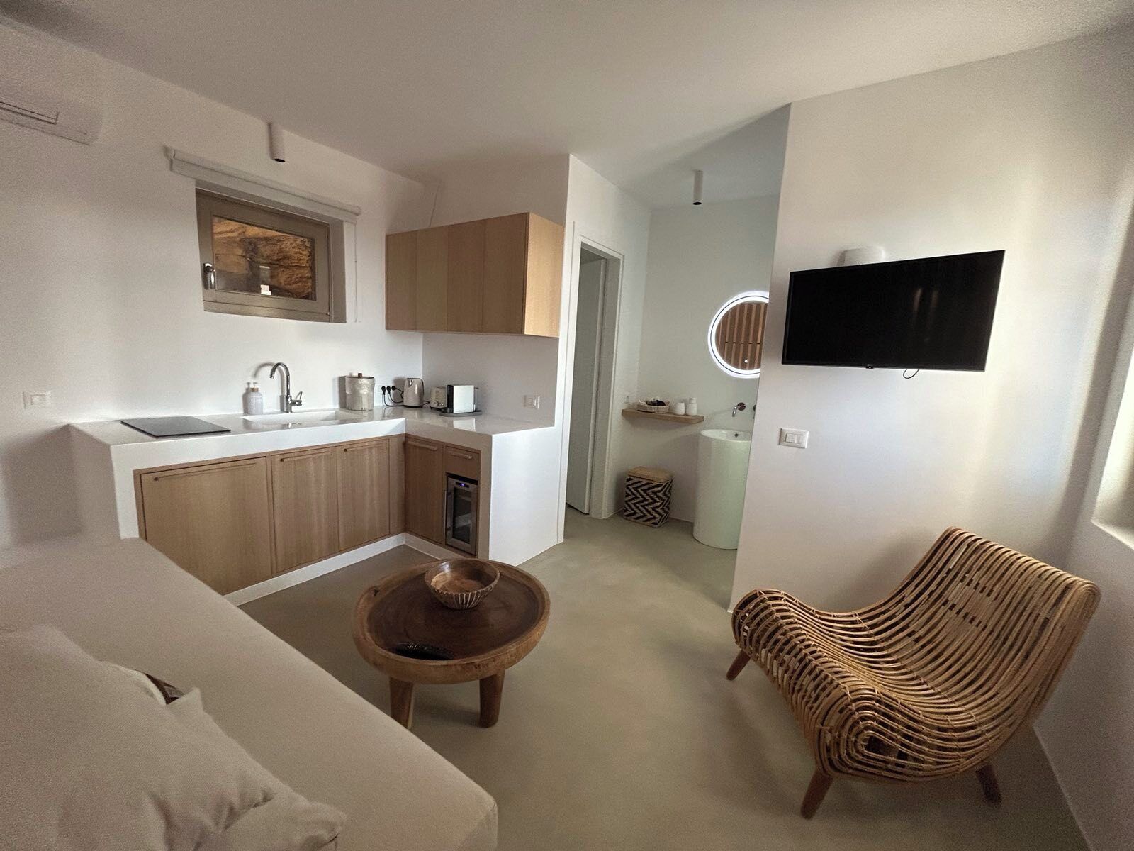 A compact living space with a kitchenette, a sofa, a round wooden table, a wicker chair, and a TV mounted on the wall.