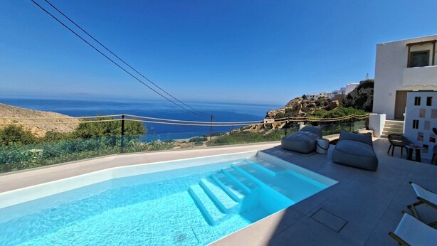 A modern outdoor pool with clear blue water, surrounded by white furniture and a scenic view of the sea and hills. - ANDELIAS SIGNATURE SUITES  (Folegandros)