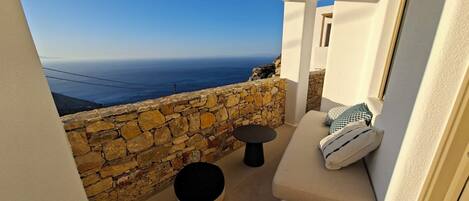 Un balcón con pared de piedra, un taburete redondo tejido, un sofá con cojines y vista al mar.